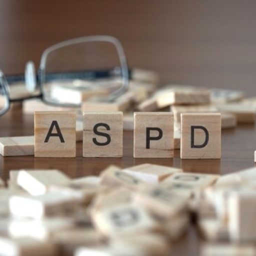Close up of wooden letter tiles spelling ASPD on a wooden surface, with additional scattered tiles and eyeglasses slightly out of focus behind them, suggesting a thoughtful or educational context.