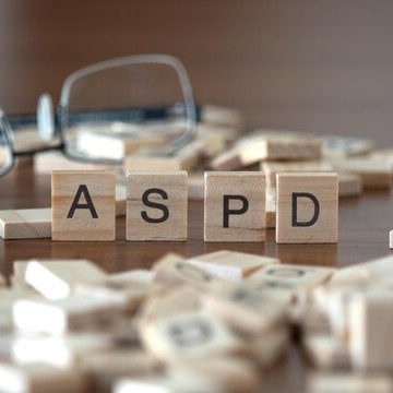 Close up of wooden letter tiles spelling ASPD on a wooden surface, with additional scattered tiles and eyeglasses slightly out of focus behind them, suggesting a thoughtful or educational context.